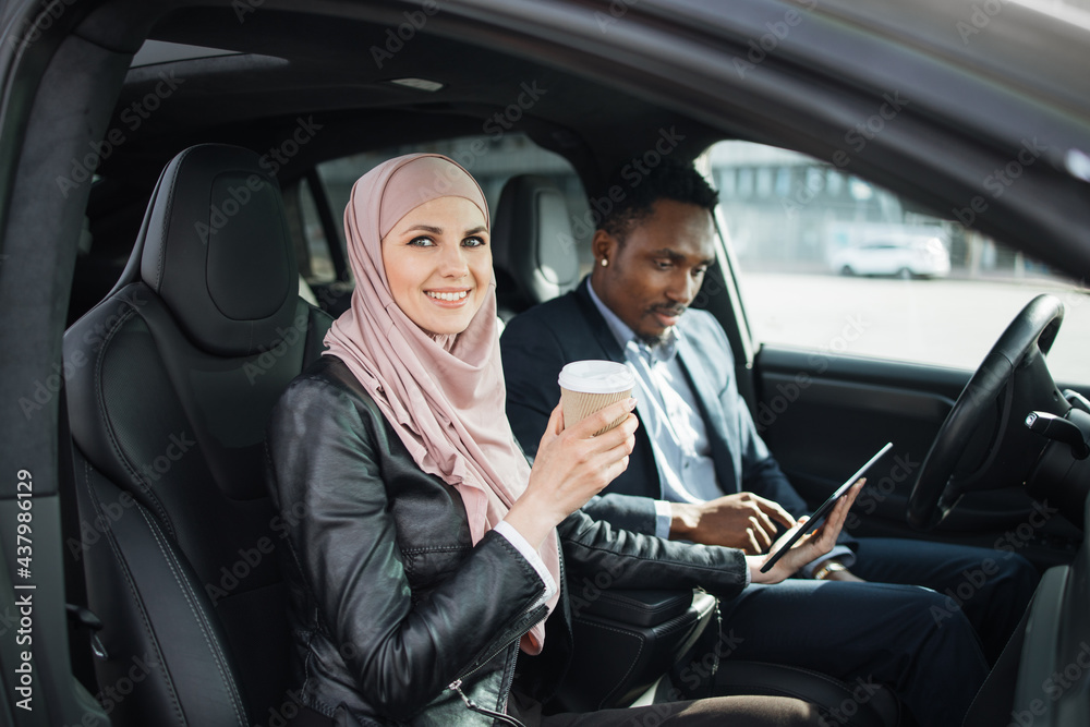 Muslim woman in hijab and african man in suit sitting together in luxury car and using digital tablet for work. Two business partners talking about common project.