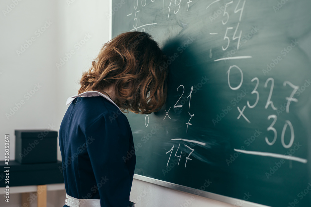 Sad girl pupil trying to solve an example. Schoolgirl stands with her forehead on the blackboard
